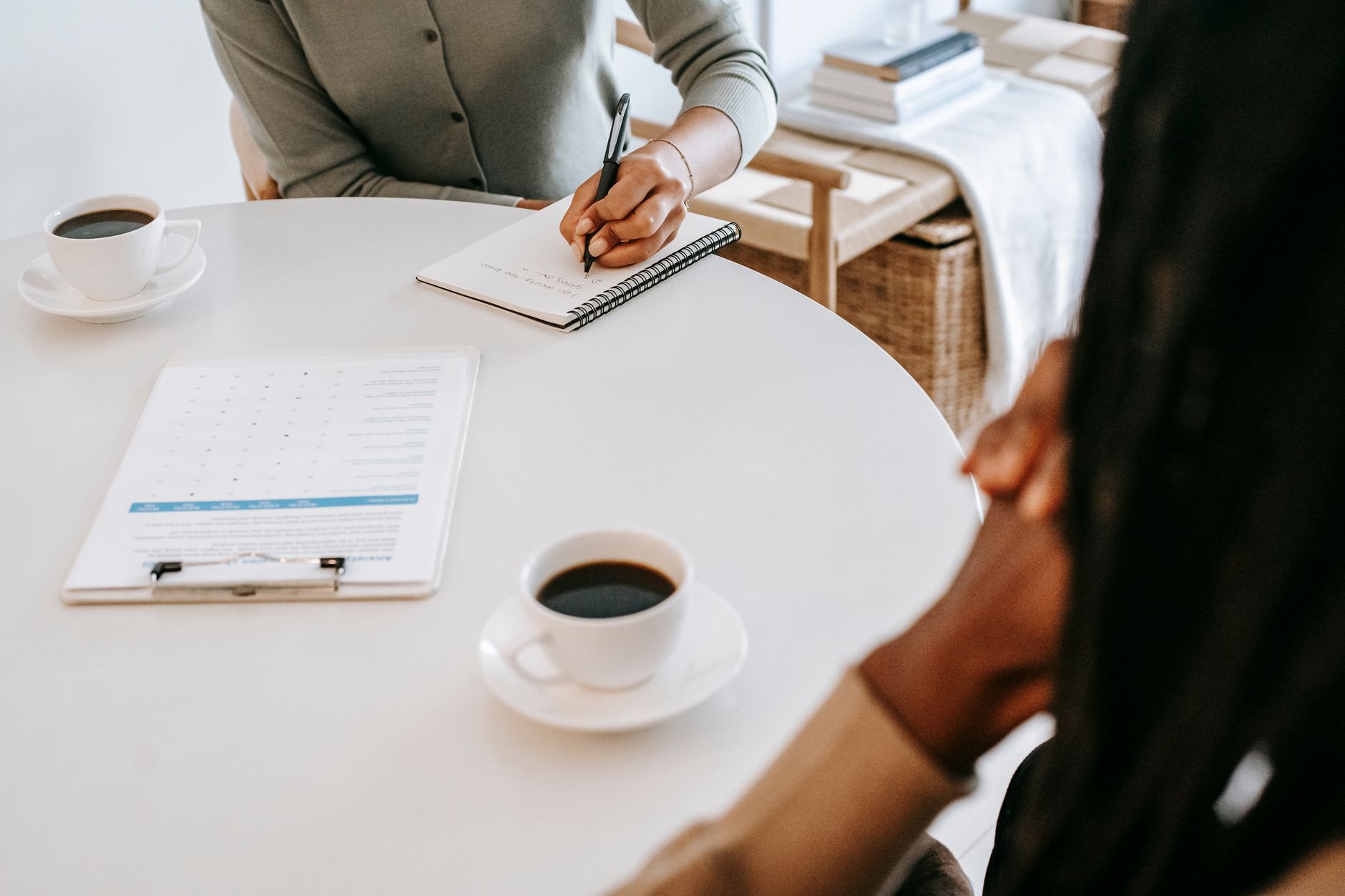 Over-the-shoulder view of a practitioner taking notes in a spiral notebook during a meeting at a round table with coffee cups and an intake form