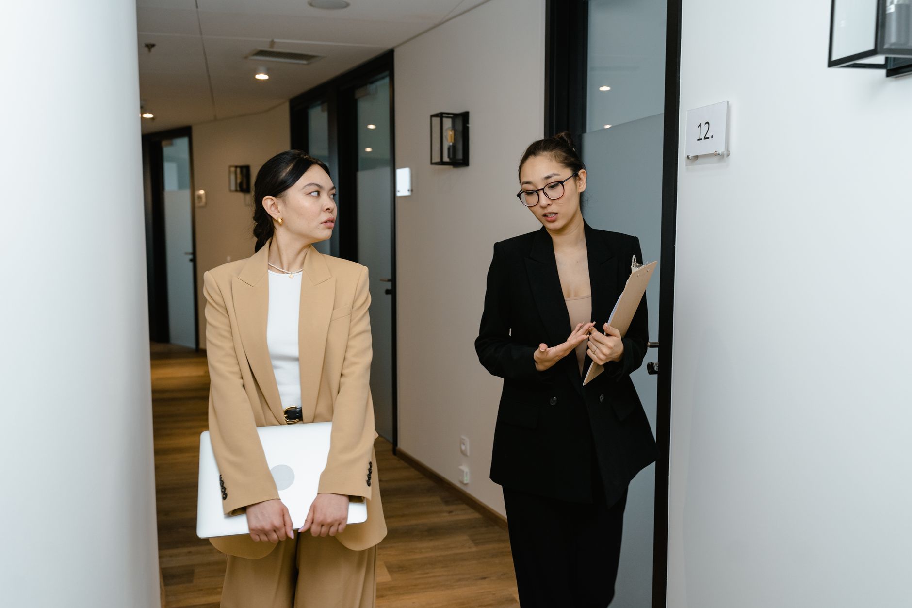 Two women in business attire talk in an office hallway, one holding a clipboard and the other a laptop