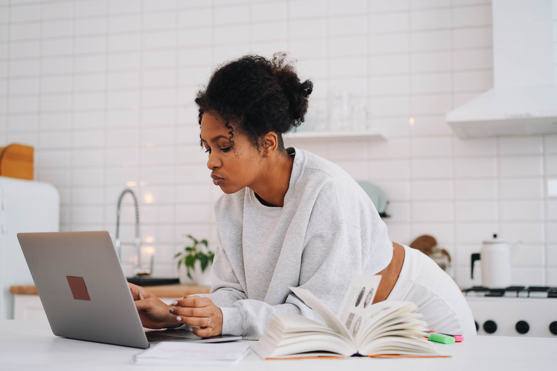 A woman leans over a kitchen counter typing on a laptop beside an open textbook and highlighters