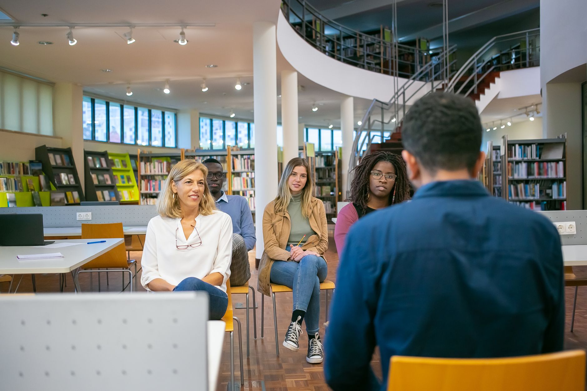 Four adult students of different ages sit in a university library and listen to a classmate speaking