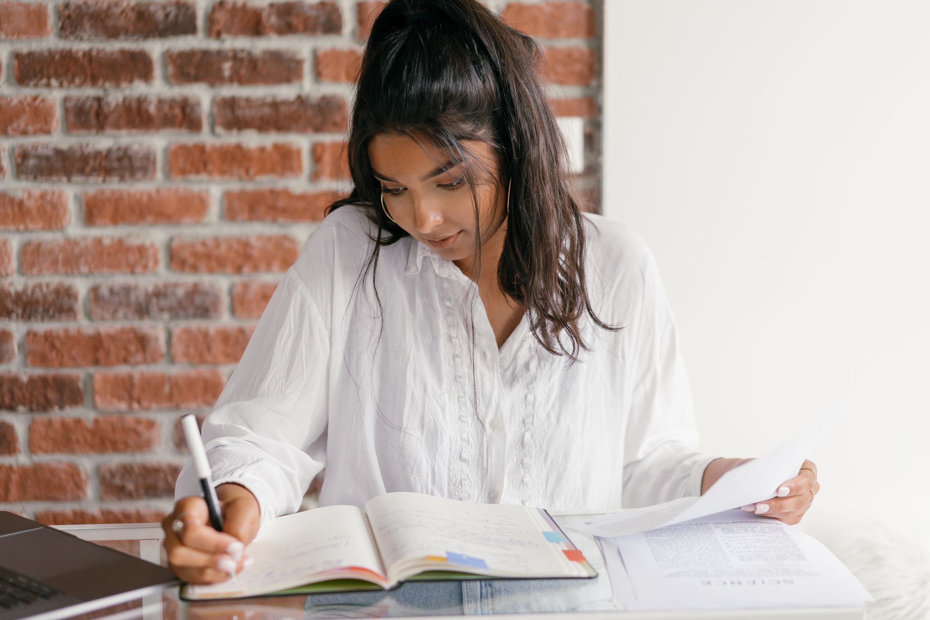 A woman in a white blouse takes handwritten notes in an open notebook beside a stack of printed pages at a desk against a brick wall