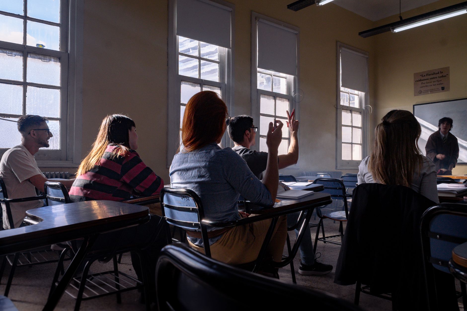 Graduate students seated at classroom desks by tall windows while one raises a hand to ask the instructor a question