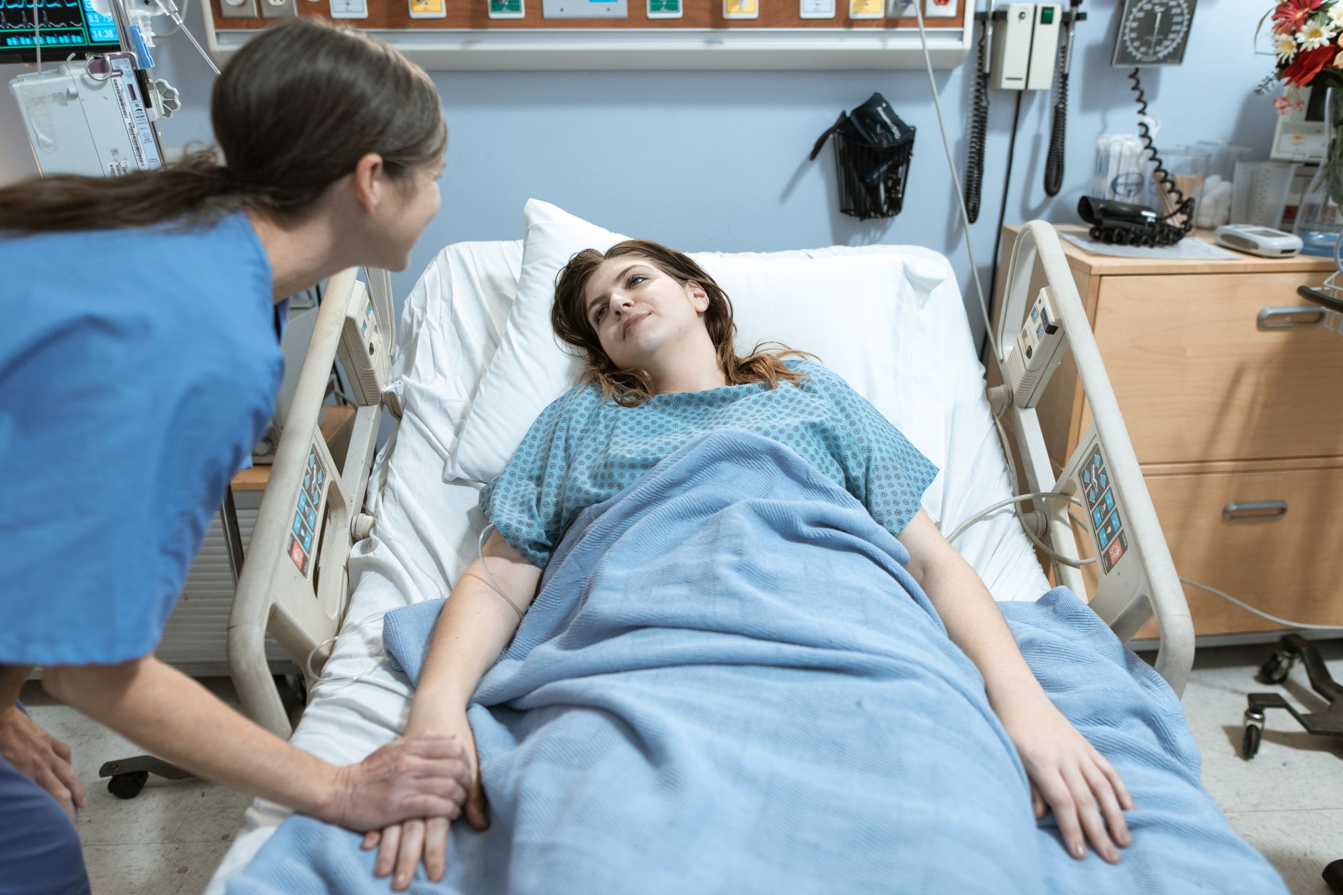 A hospital staff member leans in and holds the hand of a patient resting in a hospital bed