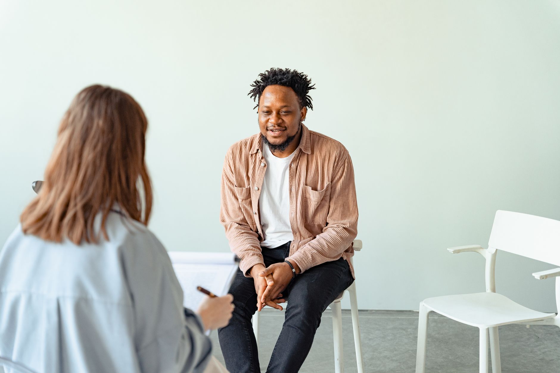 A social worker with a clipboard sits across from a smiling man in a corduroy shirt during a one-on-one meeting in a bright room