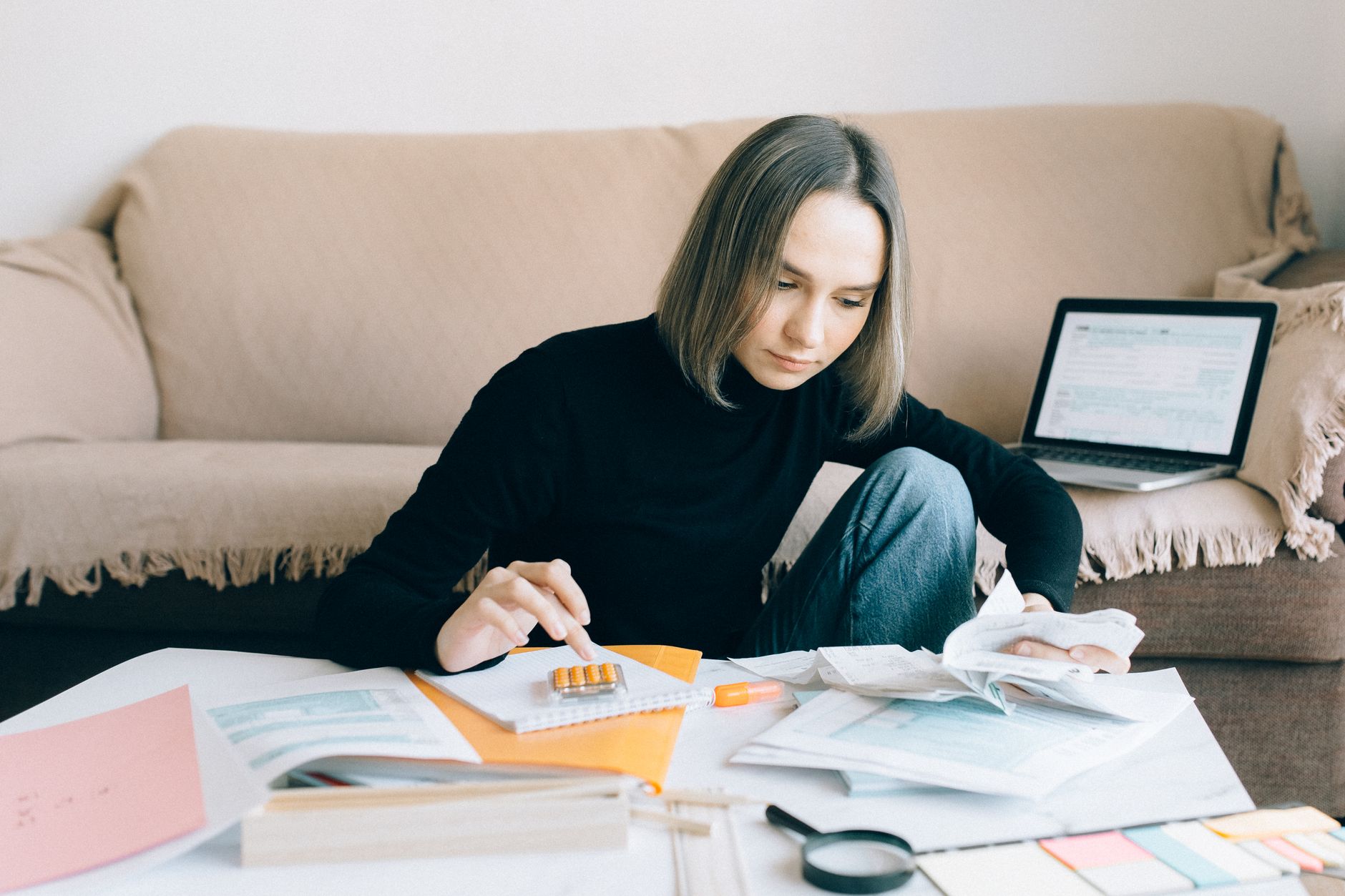 A woman sitting on the floor by a sofa, working through bills and forms with a calculator and a laptop