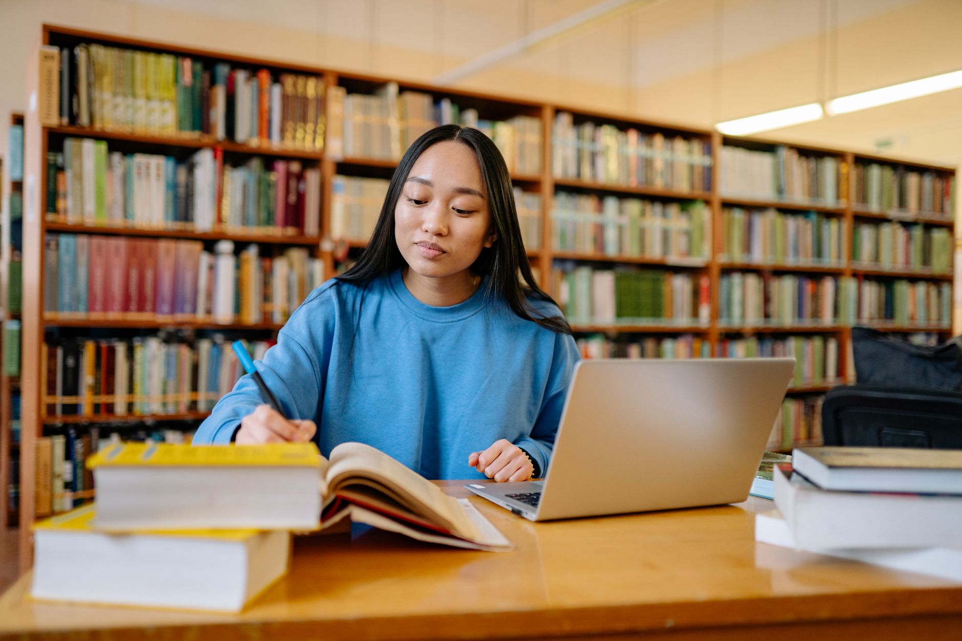 A graduate student writing notes from an open textbook beside a laptop at a library table
