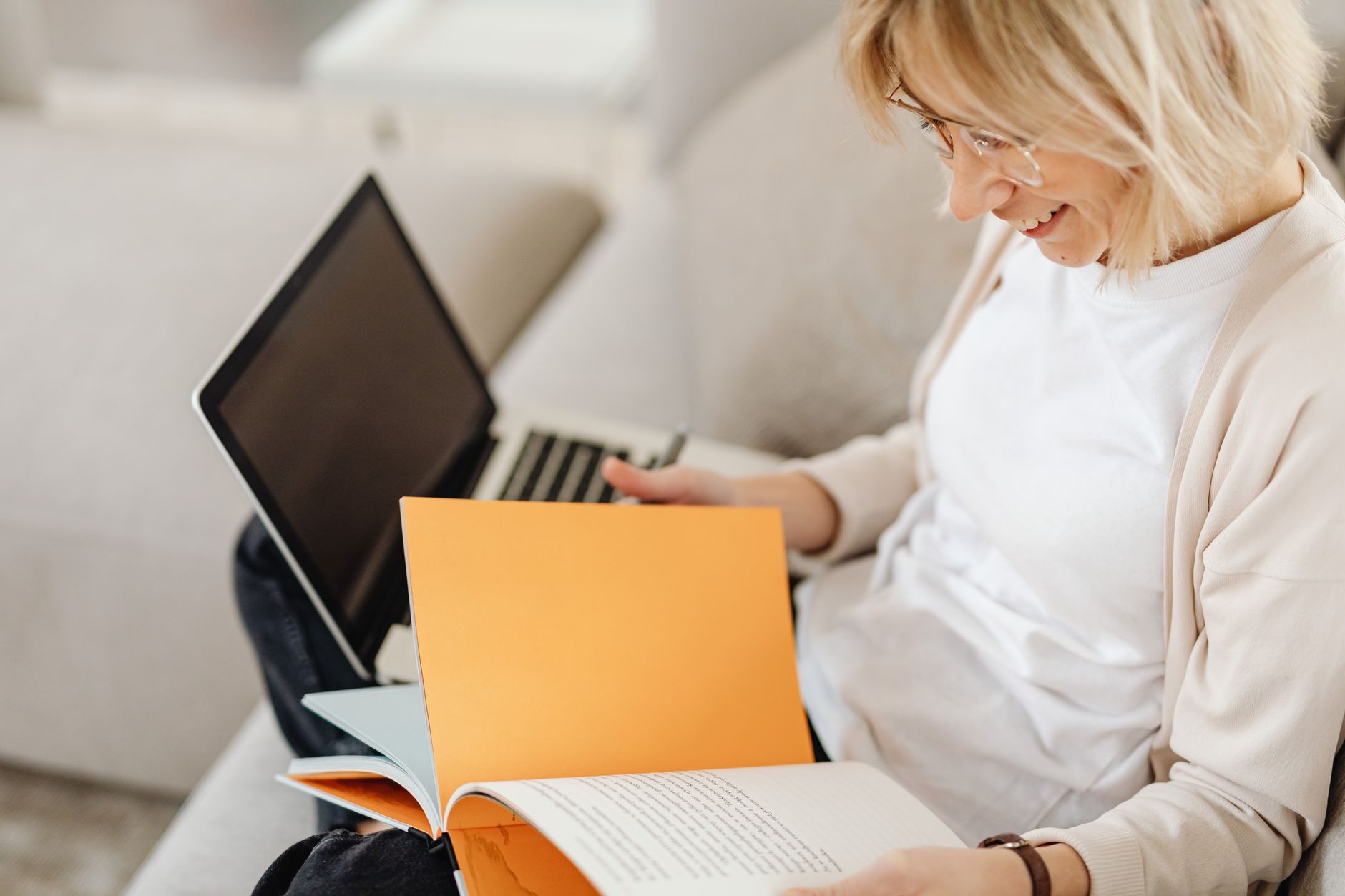 A woman with glasses reads a notebook on a sofa with an open laptop beside her