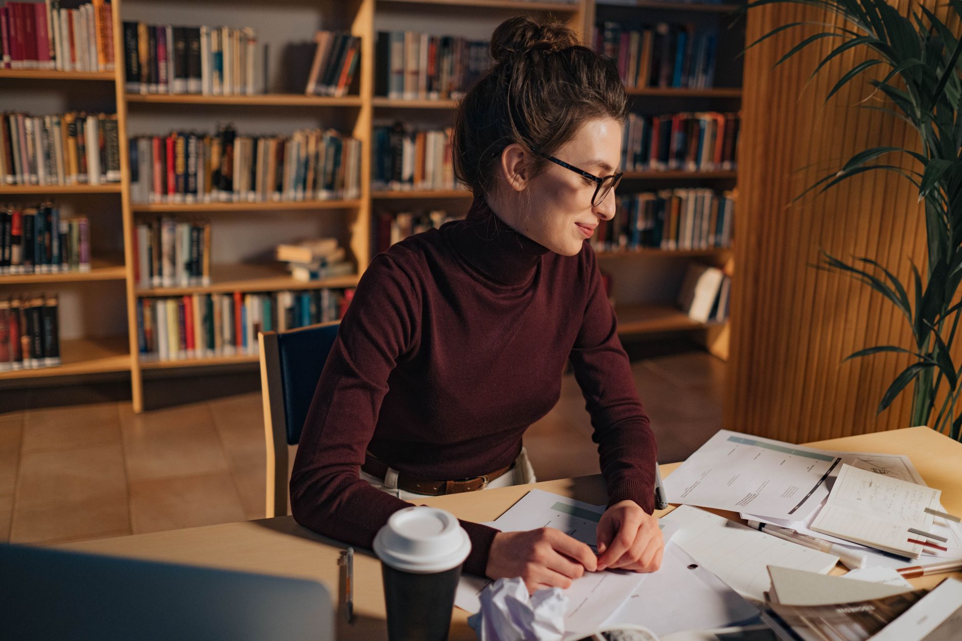 A student in glasses works through notes and papers at a library table in front of tall bookshelves