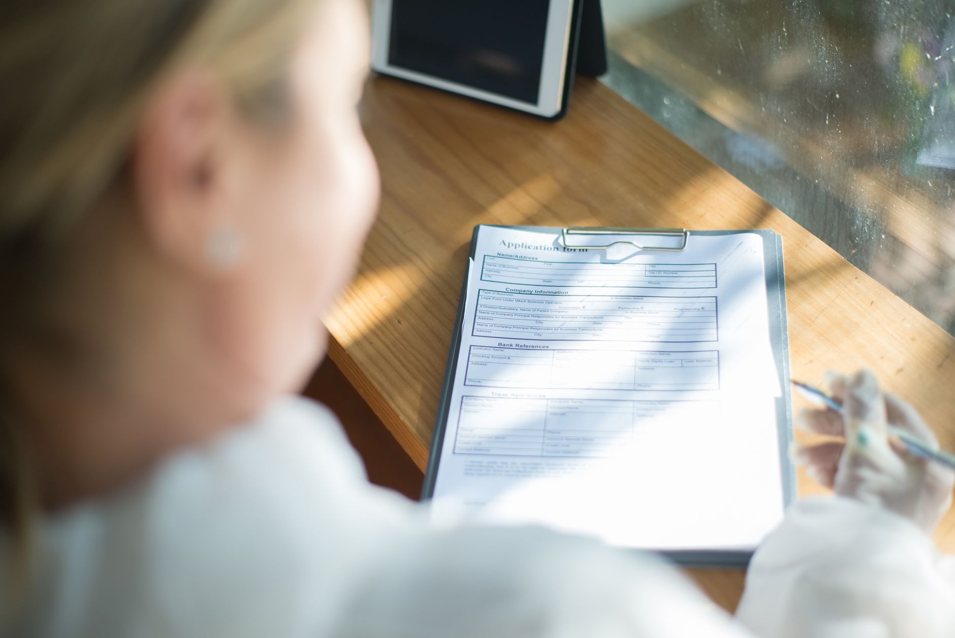 A woman with a pen works through a paper application form on a clipboard at a sunlit wooden desk beside a tablet